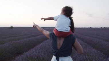 Omuzlarında küçük bir çocuk olan, mor lavanta çiçekleriyle çiçek açan ve günbatımının tadını çıkaran, tanınamayan, sevgi dolu bir annenin arka görünüşü.