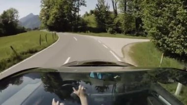 Güneşli yaz gününde, dağ vadisinde yeşil tarlalar ve ormanlar boyunca uzanan eğri asfalt yolda giden arabanın ön camından manzarayı izliyoruz.