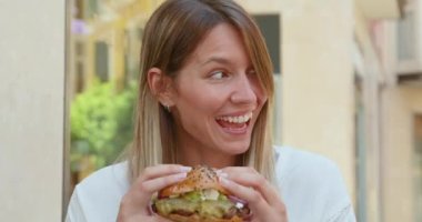 Beyaz gömlekli neşeli yetişkin bir kadın yemek için sulu burger yerken caddedeki kafe masasında arkadaşına gülümsüyor.