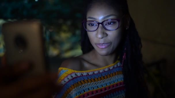 アフリカ系アメリカ人女性とビデオチャットで話している間にスマートフォン経由で暗い夜に通りに座って友人とチャット — ストック動画