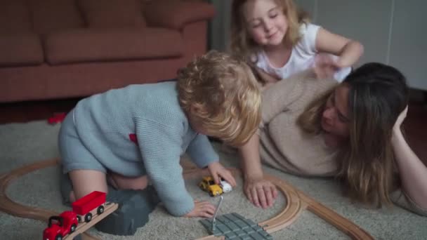 年轻女子带着小儿子和女儿在家里消磨时光的同时 还玩着玩具路和地毯上的汽车 — 图库视频影像