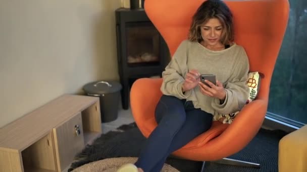 若い女性リラックスでアームチェアと自宅で携帯電話でメッセージ — ストック動画