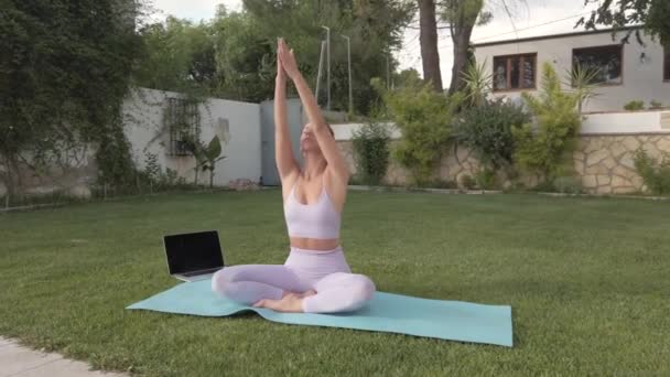クラスのためのコンピュータを使用して芝生の上でヨガを瞑想し 練習しながら 名前のジェスチャーでパドマサーナに座ってアクティブウェアの平和的な女性 — ストック動画
