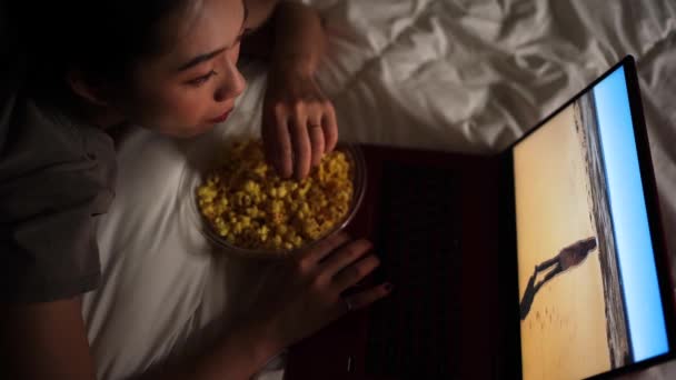 夜の映画を見ている間にポップコーンのボウルとベッドの上に横たわっているパジャマで幸せな民族の女性の上から — ストック動画