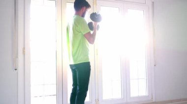 Spor kıyafetleri içinde çıplak ayaklı bir erkek sporcunun parlak mavi bir spor minderinin üzerinde, evdeki geniş ışıklı odada halterle egzersiz yaparken yan görüntüsü.