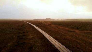 İzlanda 'da gün batımında kuru tarlalardan dağlara doğru giden dar asfalt yolda drone görüntüsünü yakınlaştır