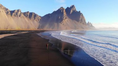 Stoksnes, İzlanda 'daki dağların yakınında deniz kıyısında yürüyen tanınmayan bir gezginin hava manzarası.