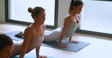 Spor kıyafetleri içinde, yoga sırasında karın kaslarını esneten formda kadın sporcuların geniş bir stüdyoda yukarı baktıkları yan manzara.