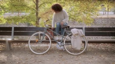 Kıvırcık saçlı, sonbahar bahçesinde bisikletin yanında bankta oturan ve hafta sonları akıllı telefondan arkadaşlarıyla mesajlaşan Afro-Amerikan kadın.