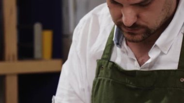 Çağdaş atölyenin bulanık iç mekânına karşı masada özel giyim koleksiyonu üzerinde çalışırken gelecekteki kıyafetlerin detaylarını inceleyen ya da geliştirenler üzerine kalemle yazılar yazan kırpıcı genç erkek terzi.