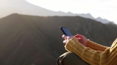 Güneşli bir günde dağ sırasının arka planında otururken cep telefonuyla mesajlaşan pozitif kadın kaşifin yan görüntüsü