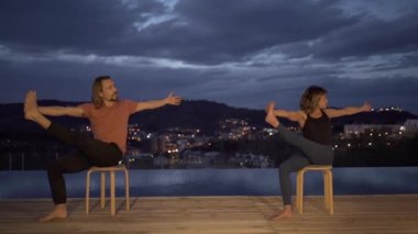 Aktif kıyafetli sakin bir adam ve kadın gece boyunca yüzme havuzunun yanındaki terasta birlikte farkındalık çalışırken yoga yapıyorlar.