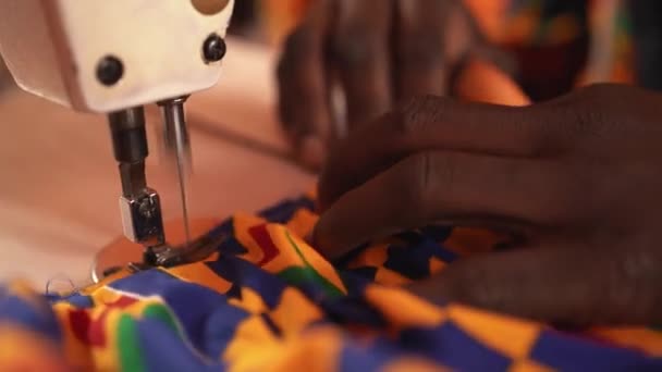 裁缝机上用非洲图案的彩色面料做国家服装的作物男裁缝 同时坐在非洲彩色面料和缝纫车间的模特背景上 — 图库视频影像