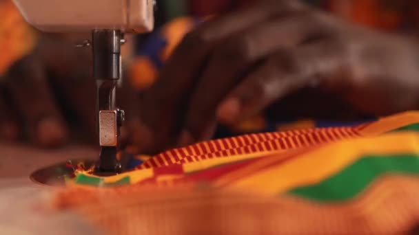 縫製工場で着色されたアフリカの材料とマネキンを背景に座っている間 ミシンにアフリカのパターンを持つ色の布の民族衣装を作成するクロップ男性の仕立て — ストック動画