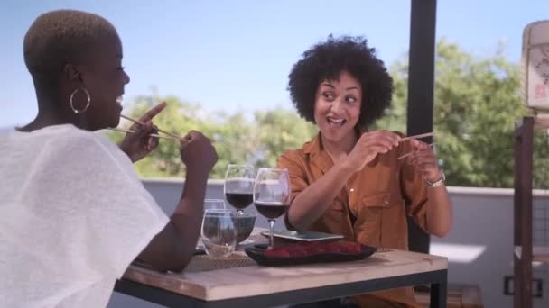 头发卷曲的黑人妇女解释了非洲裔美国女性朋友在咖啡桌旁等待点菜时如何使用筷子 — 图库视频影像