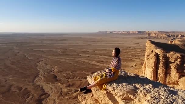 在多风的天气里 身穿黄色衣服的女游客坐在陡峭的岩石悬崖边 在无云的天空中欣赏干旱的沙漠 — 图库视频影像