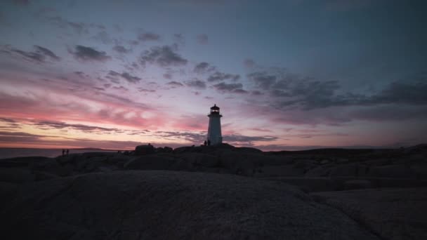 カナダの日没時に波打つ海の近くに位置するPeggys Cove Lighthouseで岩の多い海岸を賞賛する観光客 — ストック動画