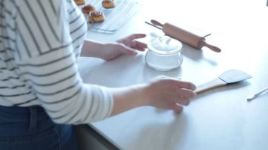Kimliği belirsiz bir kadının, sıcak mutfaktaki tatlı ruloların yanındaki beyaz masadan şeker ve spatula aldığı yüksek açı.