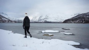 Ilık ceketli ve şapkalı genç Asyalı kadın etrafta dolanıyor ve sonra diz çöküp beyaz ve kahverengi karlı tepelere ve Norveç 'teki Kaldfjorden gölünün sakin serin suyuna yaslanıyor.