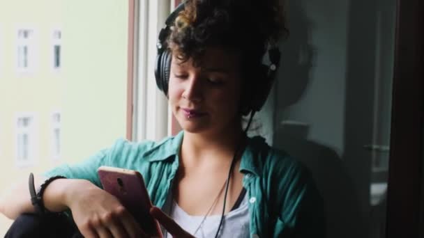 自宅でスマートフォンを使用してヘッドフォンで陽気な女性 — ストック動画