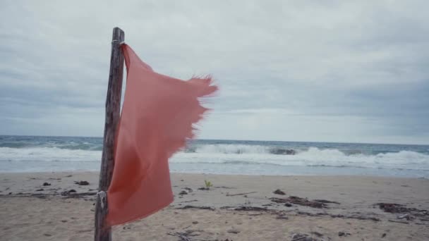 在空旷的沙滩上挂着摇曳着红布的木棍 背景是汹涌的海浪 — 图库视频影像