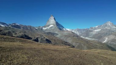 Matterhorn dağının görkemli manzarası berrak mavi gökyüzünün altında, engebeli araziyle çevrili. Manzaranın üstündeki ikonik tepe kuleleri ihtişamını ve doğal güzelliğini gözler önüne seriyor..