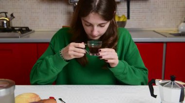 Mutfak masasında oturan genç bir kadın serinletmek için bir fincan sıcak kahve üflüyor. Yeşil bir kazak giyiyor ve sakin ve rahat görünüyor. Evde sakin bir sabah anının tadını çıkarıyor.