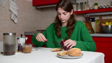 Bir kadın, sabah kahvaltısını rahat bir mutfakta hazırlar, taze malzemeler ve sıcak bir ortam ile çevrili, gevşemiş ve odaklanmış bir tavırla ekmeğe reçel sürer..
