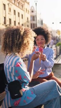 İki arkadaş şehir merkezindeki bir bankta oturmuş güneşli bir günde dondurmanın tadını çıkarıyorlar. Gülüşleri ve gülüşleri şehirde paylaşılan neşeli bir anı yansıtıyor..