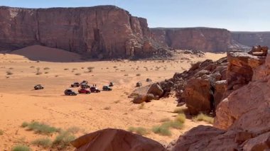 Bir grup off-road aracı, açık mavi gökyüzünün altındaki yüksek ve kayalık uçurumlarla çevrili Libya 'nın Akakus kentinin engin ve manzaralı çöl manzarasında toplandı.