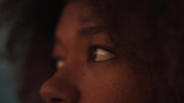 Yumuşak, kıvırcık saçlarla çerçevelenmiş, mesafeye bakan düşünceli gözleri gösteren yakın plan bir görüntü. Işıklandırma, doğal güzelliği vurgulayan sıcak ve samimi bir atmosfer yaratıyor..