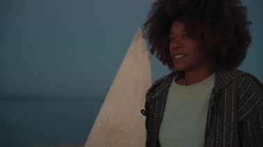 Taghazout, Fas 'ta sakin bir günde bir kadın sörf tahtasının yanında duruyor. Huzurlu okyanus ileriye doğru uzanıyor, barışçıl ve yansıtıcı bir atmosfer yaratıyor..