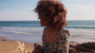 Kıvırcık saçlı bir kadın Taghazout, Fas 'ın kumlu sahillerinde güneşli bir günün tadını çıkarır. Açık mavi gökyüzü ve yumuşak dalgalar dingin ve resimli bir plaj sahnesi yaratır..