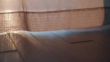 Bir kadının ayaklarının yakın çekimi, yüksek topuklu ayakkabılar içinde, perdeden süzülen yumuşak, sıcak bir ışıkla aydınlatılmış ahşap bir zeminde. Samimi ve sinematik atmosfer.