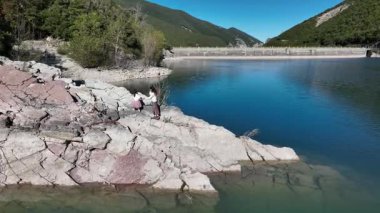 İtalya 'da kayalık bir göl kıyısında yürürken kızının elini tutan bir kadının hava görüntüsü. Sakin manzarada berrak mavi su ve yemyeşil alanlar var..