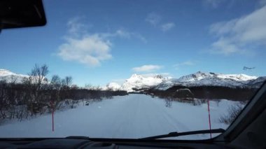 Açık mavi bir kış gününde Norveç 'in Lofoten kentindeki yapraksız ağaçlar ve dramatik dağlar arasındaki mesafeye uzanan karla kaplı bir yol..