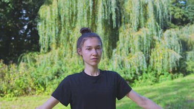 Bir kadın açık havada yoga yapıyor, gözleri kapalı ve kolları havada duruyor, yemyeşil doğal bir ortamda huzur ve farkındalığı somutlaştırıyor, rahatlığı ve sağlığı teşvik ediyor..