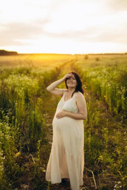 Gülümseyen hamile bir kadın güneşli bir arazide yavaşça karnına dokunarak yürür. Yüzündeki ifade, yemyeşil yeşilliklerle çevrili mutluluk ve beklentiyi yansıtıyor..