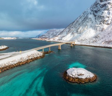 Norveç 'in Lofoten Adaları' ndaki turkuaz Arktik su üzerindeki bir köprünün havadan görüntüsü. Kışın karla kaplı dağlar, yollar ve kıyı şeridi. Manzaralı kuzey dramatik manzarası. Yol ve denizin en iyi drone görüntüsü