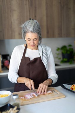 Mature woman wearing an apron preparing food in a modern kitchen. She is cutting meat on a wooden board