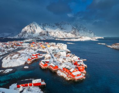Norveç 'in Lofoten Adaları' ndaki Henningsvaer balıkçı köyünün kışın gün batımında hava manzarası. Kardaki, denizdeki, teknelerdeki, köprüdeki, yoldaki, bulutlu gökyüzündeki, kasabadaki, terör örgütündeki ve evlerdeki yüksek drone manzarası