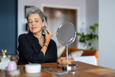 Olgun bir kadın yüzüne gua sha taşıyla masaj yapıyor. Ev ortamında cilt gençleştirme ve rahatlatmayı teşvik ediyor.
