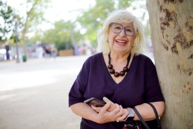 Kıdemli kadın gülümsüyor ve elinde akıllı bir telefonla parkta bir ağaca yaslanıyor.