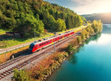 Sonbaharda gündoğumunda Alp Dağları 'nda nehir kıyısında hareket eden kırmızı, modern hızlı trenin havadan görünüşü. Trenin üst manzarası, kırsal demiryolu, göl, yol, sonbaharda yeşil ağaçlar. Slovenya 'daki tren istasyonu