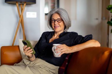 Gülümseyen olgun bir kadın kanepede dinleniyor, akıllı telefonunu kullanıyor ve bir fincan kahve içiyor.