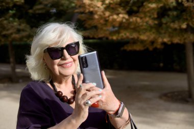 Parkta akıllı telefonuyla fotoğraf çeken, teknoloji ve açık hava eğlencesinin tadını çıkaran, güler yüzlü son sınıf öğrencisi bir kadın.