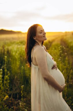 Hamile bir kadın güneşli bir alanda kameraya hafifçe gülümsüyor. İki eliyle göbeğini kucaklıyor, yemyeşil ve ılık doğal ışıkla çevrili..