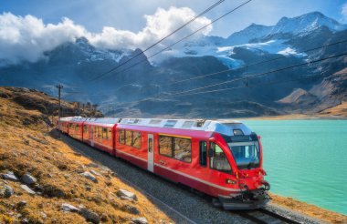 Sonbaharda İsviçre 'de güneşli bir sonbahar gününde İsviçre alp dağlarında demiryolu boyunca ilerliyorlar. Bernina Ekspres treni, turuncu çimen, bulutlardaki karlı kayalar, sonbaharda günbatımında mavi gökyüzü