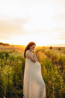 Hamile bir kadının gün batımında güneşli bir çayırda dikilirken yan görünüşü anneliği, sükuneti ve yeni bir hayat beklentisini temsil ediyor..