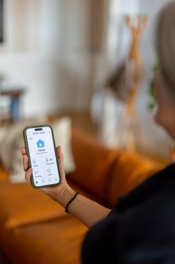 Akıllı telefon tutan ve mobil uygulama, kontrol ışıkları, sıcaklık ve güvenlik sistemlerini kullanarak akıllı ev aletlerini yöneten kadın